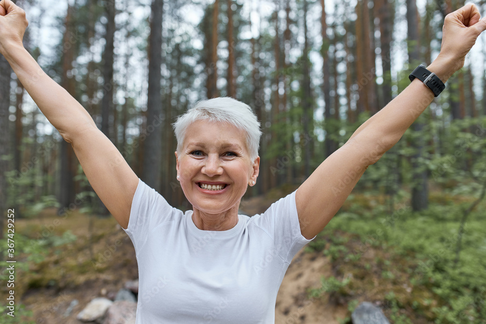 Close up view of energetic confident middle aged woman runner raising hands, rejoicing at success as she broke her own record, having excited facial expression. Happy sporty female posing outdoors Close up view of energetic confident middle aged woman runner raising hands, rejoicing at success as she broke her own record, having excited facial expression. Happy sporty female posing outdoors