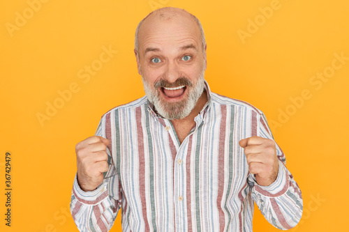 Success, celebration, excitement and victory concept. Attractive cheerful senior man with thick beard clenching fists and screaming Yes, celebrating good poositive news, having overjoyed ecstatic look photo