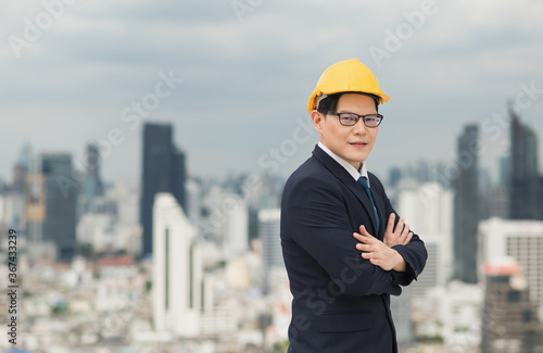 Young handsome asian civil engineer standing with arms crossed wearing helmet yellow hard hat in business suite on building urban cities background.