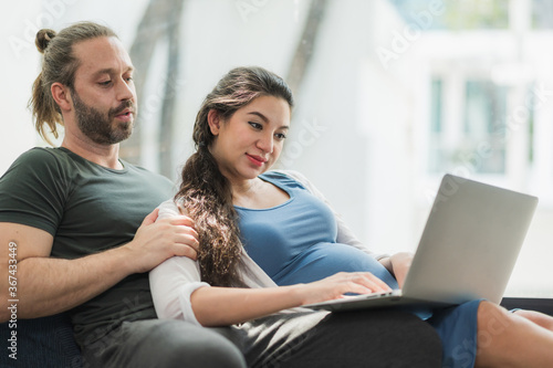 Beard man and his pregnant wife watching movie on laptop feeling happy while sitting on sofa in the home. Family concept. © Nopphon