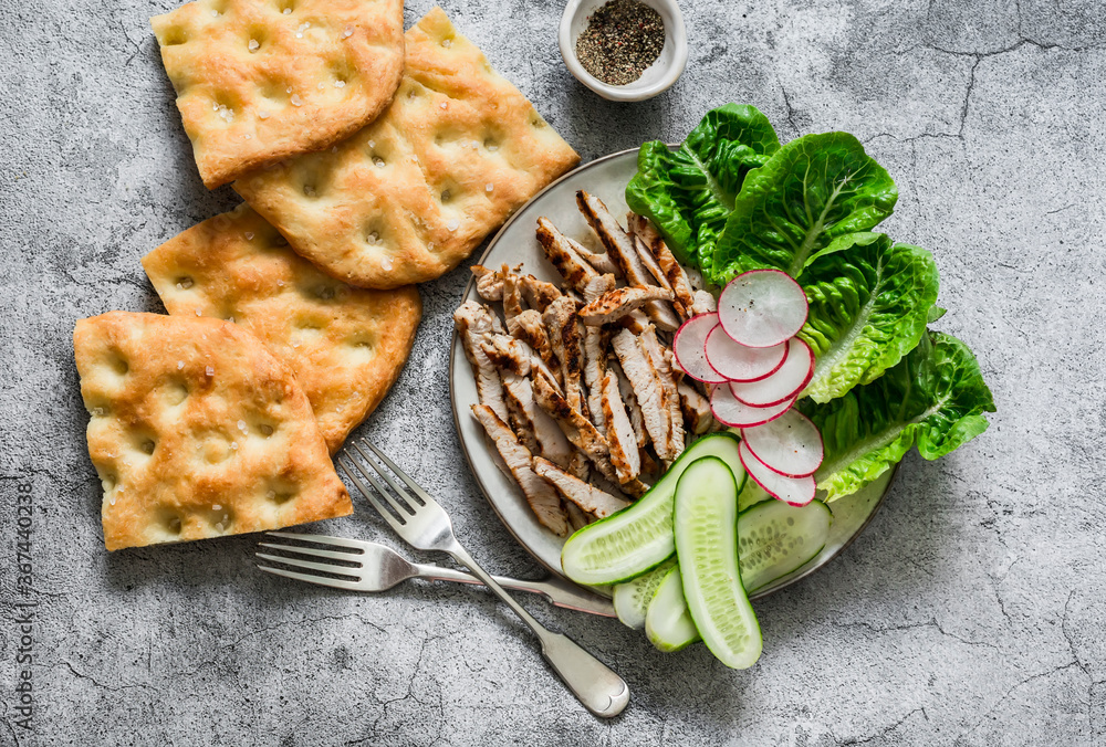 Mediterranean lunch - crispy italian focaccia, grilled turkey, green romaine salad and fresh vegetables on a grey background, top view