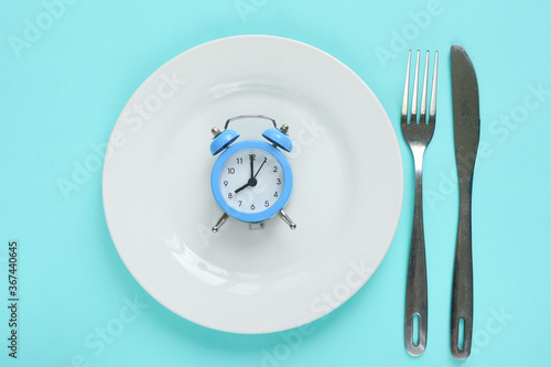 Blue alarm clock, fork, knife and empty plate on colored paper background. Intermittent fasting concept- Image. Time concept. Breakfast concepte.