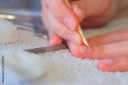 Woman pushing cuticle using wooden stick on her nail making manicure herself at home, hands closeup. Hygiene and care about nails. Beauty procedure. Tools and cotton pads on towel on table.