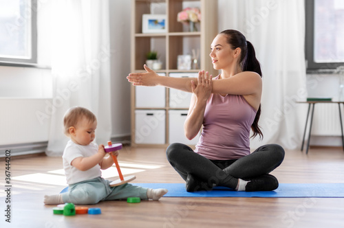 family  sport and motherhood concept - happy smiling mother exercising on mat and little baby playing with toys at home