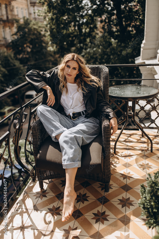 Stylish fashionable blonde woman with smoky eye makeup, in jeans, white T-shirt and black leather jacket on the balcony in an armchair. Spring autumn fashion concept. Soft selective focus.