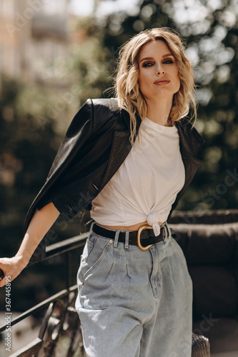 Stylish fashionable blonde woman with smoky eye makeup, in jeans, white t-shirt and black leather jacket on the balcony in the city. Spring autumn fashion concept. Soft selective focus.