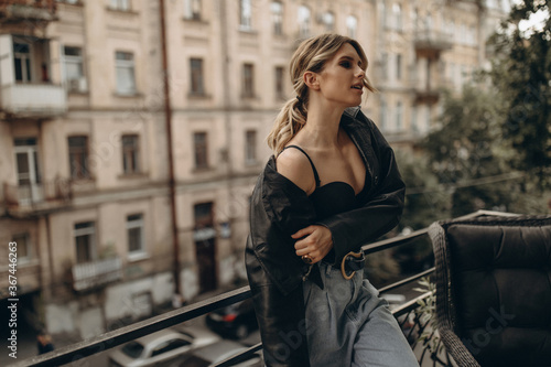 Stylish fashionable blonde woman with smoky eye make-up, in jeans, lingerie and black leather jacket dancing on a balcony in the city. Spring autumn fashion concept. Soft selective focus.