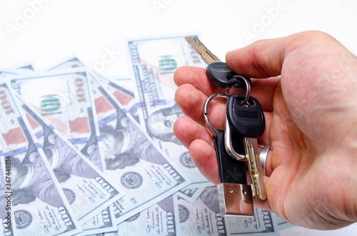 Two black keys with a leather keychain in close-up lie in a man's hand on the background dollars. Buying, selling real estate, apartment, house, car, business