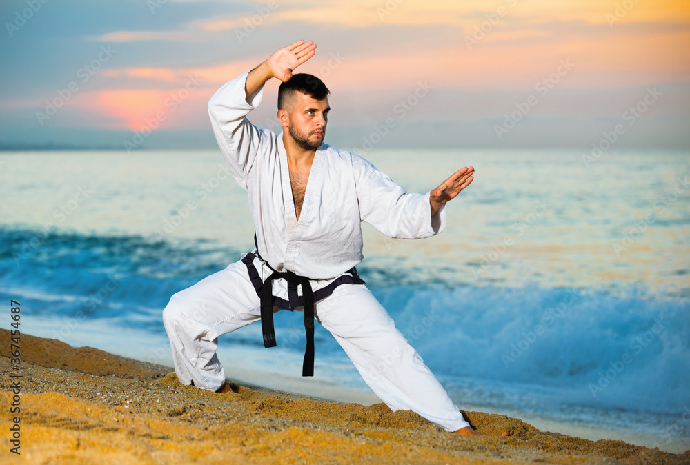 Friendly smiling pleasant man in uniform doing taekwondo exercises at sunset sea shore
