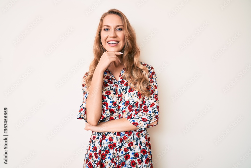 Young caucasian woman with blond hair wearing summer dress looking confident at the camera smiling with crossed arms and hand raised on chin. thinking positive. Young caucasian woman with blond hair wearing summer dress looking confident at the camera smiling with crossed arms and hand raised on chin. thinking positive.