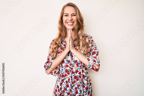 Young caucasian woman with blond hair wearing summer dress praying with hands together asking for forgiveness smiling confident.