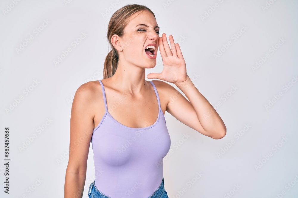 Young blonde girl wearing casual style with sleeveless shirt shouting and screaming loud to side with hand on mouth. communication concept. Young blonde girl wearing casual style with sleeveless shirt shouting and screaming loud to side with hand on mouth. communication concept.