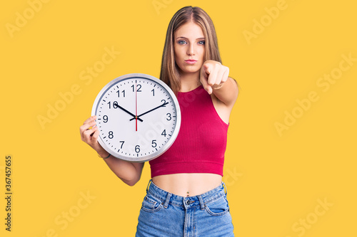 Young beautiful blonde woman holding big clock pointing with finger to the camera and to you, confident gesture looking serious