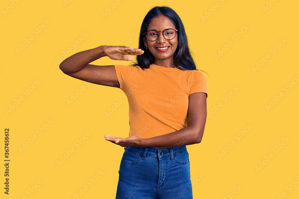 Young indian girl wearing casual clothes and glasses gesturing with hands showing big and large size sign, measure symbol. smiling looking at the camera. measuring concept. Young indian girl wearing casual clothes and glasses gesturing with hands showing big and large size sign, measure symbol. smiling looking at the camera. measuring concept.
