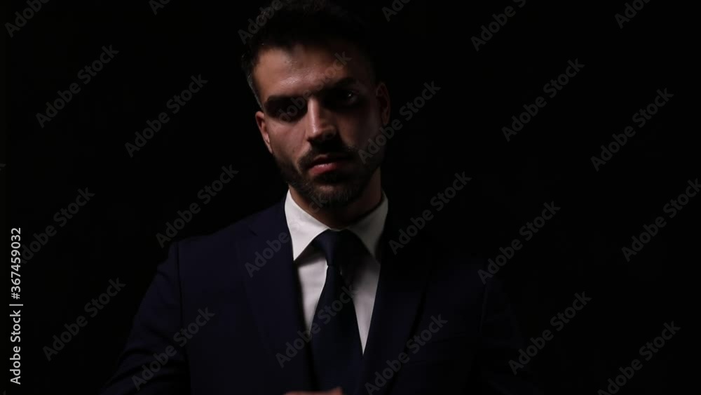 mysterious young businessman adjusting suit and tie in a shadow light, moving in a side view pose and walking away on a dark background mysterious young businessman adjusting suit and tie in a shadow light, moving in a side view pose and walking away on a dark background