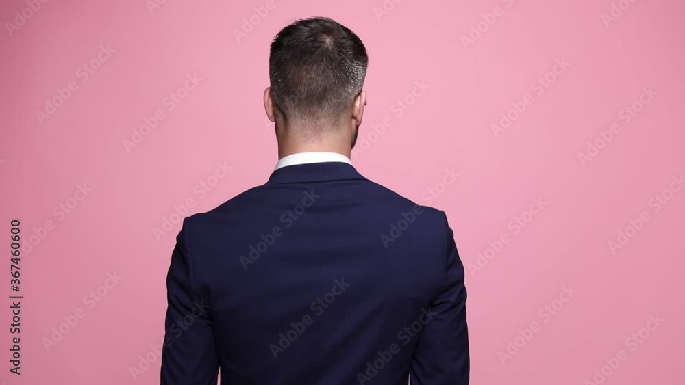 young handsome man in navy suit is standing with his back at the camera, slowly turning his head aside, wearing sunglasses and having a cool attitude on pink background young handsome man in navy suit is standing with his back at the camera, slowly turning his head aside, wearing sunglasses and having a cool attitude on pink background