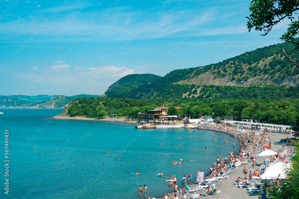 Beautiful coast of the Black Sea with a crowd of tourists on a background of green hills. The beach with many tourists resting in the sun with hot weather.