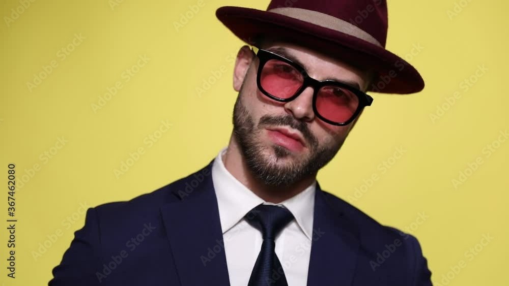 sexy businessman in suit wearing hat and sunglasses moving head in both sides, holding hand to face and touching beard and chin, thinking and admiring, flirting on yellow background sexy businessman in suit wearing hat and sunglasses moving head in both sides, holding hand to face and touching beard and chin, thinking and admiring, flirting on yellow background