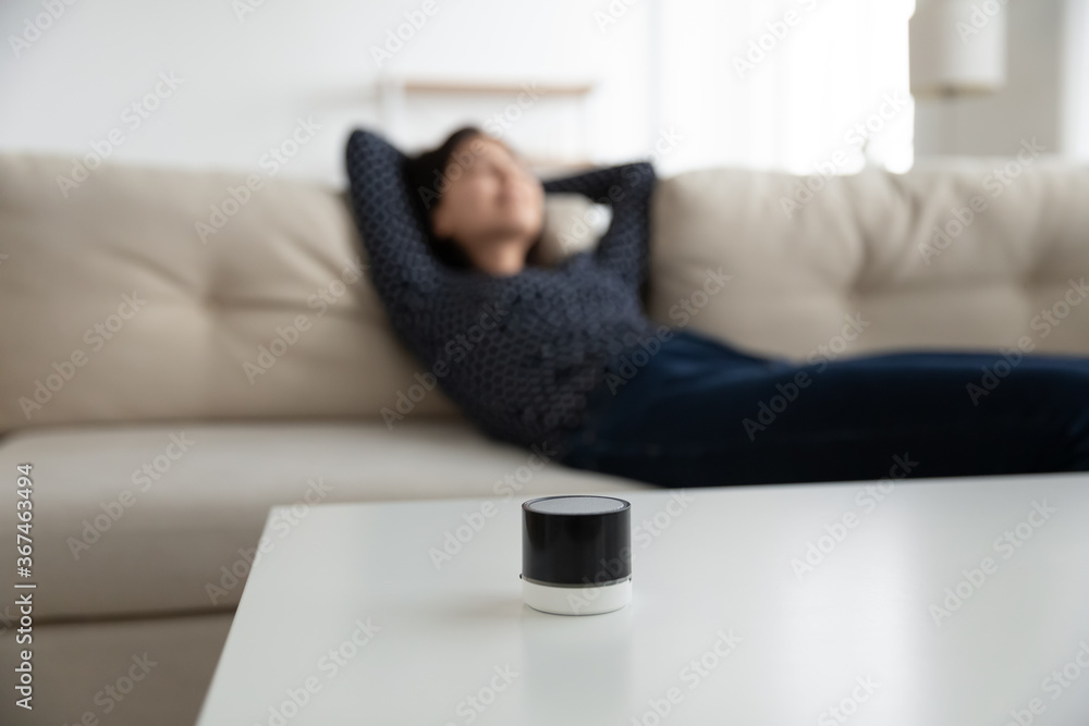 Focus on mini modern smart wireless speaker on table, woman relax rest on sofa in living room at background, female listen to music good quality sound loudspeaker audio device on couch Focus on mini modern smart wireless speaker on table, woman relax rest on sofa in living room at background, female listen to music good quality sound loudspeaker audio device on couch