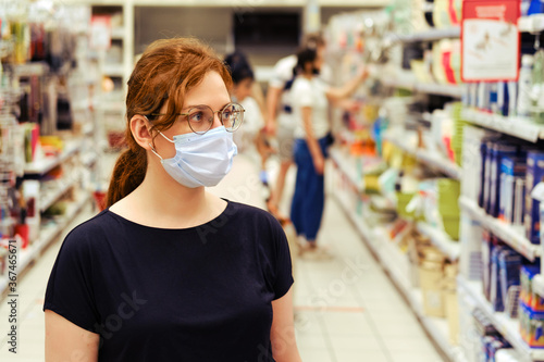 Young woman in medical mask between rows with shelves in a store. Visiting public places during the coronavirus pandemic
