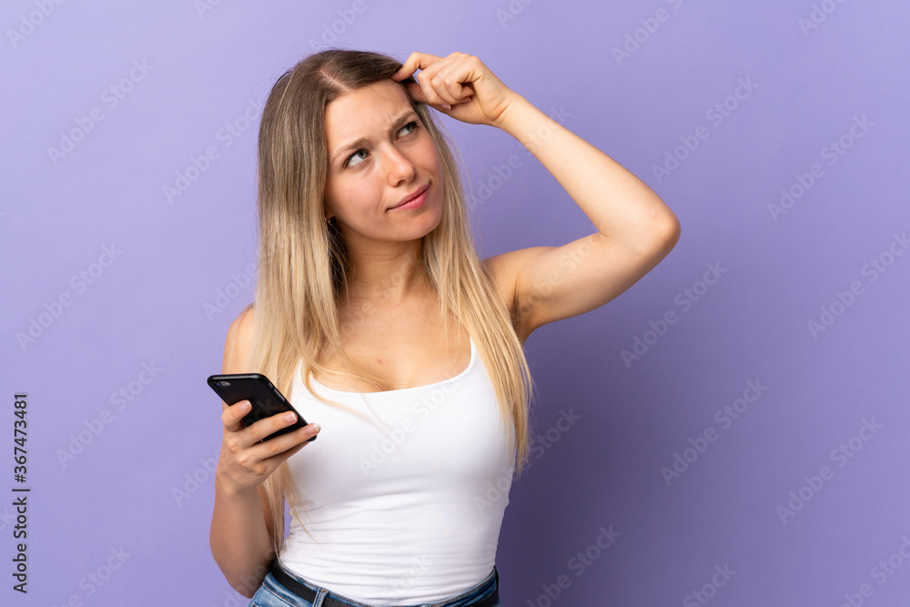 Young blonde woman using mobile phone isolated on purple background having doubts and with confuse face expression Young blonde woman using mobile phone isolated on purple background having doubts and with confuse face expression