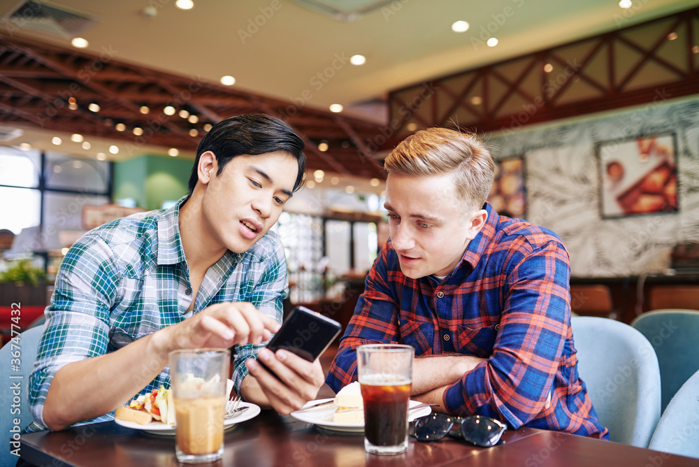 Young Asian man having lunch with friend in cafe and showing new mobile application on smartphone to friend