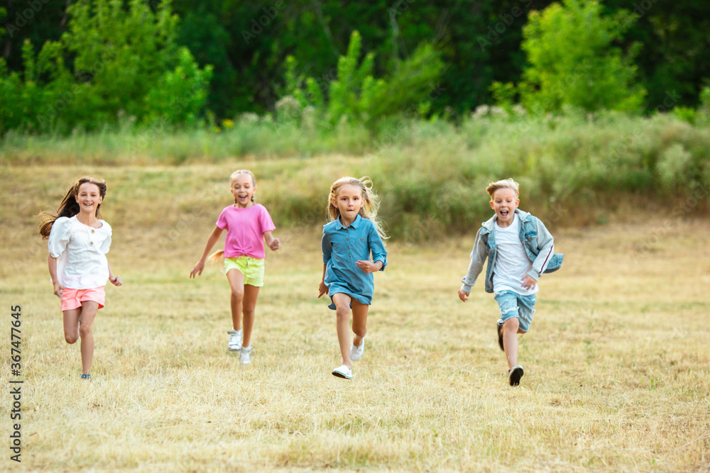 Fototapeta premium Kids, children running on meadow in summer's sunlight. Look happy, cheerful with sincere bright emotions. Cute caucasian boys and girls. Concept of childhood, happiness, movement, family and summer.