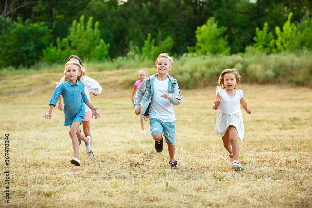 Fototapeta premium Kids, children running on meadow in summer's sunlight. Look happy, cheerful with sincere bright emotions. Cute caucasian boys and girls. Concept of childhood, happiness, movement, family and summer.
