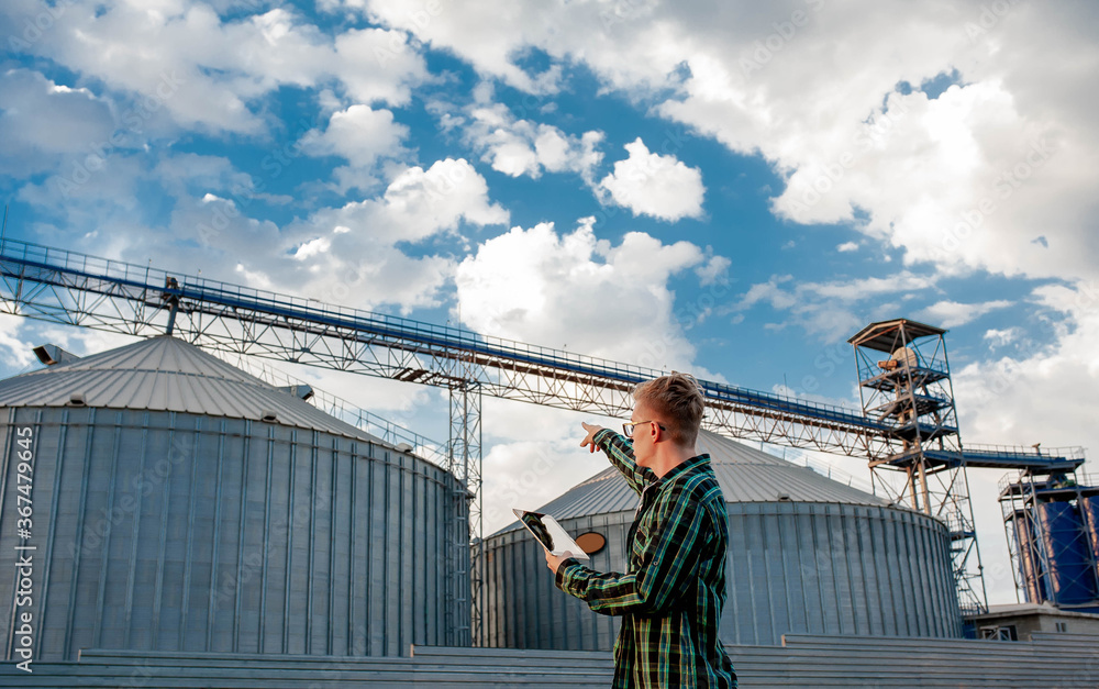 
A young guy stands with a tablet near a metal elevator in an agricultural area. A grain warehouse.