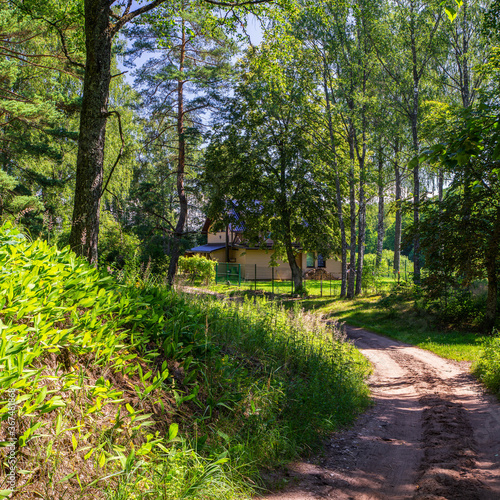 Rural dusty road between green trees to the countryside private house. Modern exterior. Siding wall. Summer landscape.