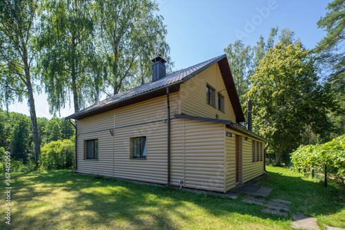 Countryside private house near the green forest. Modern exterior of cottage. Siding walls with windows. Tiled roof with chimney pipe.