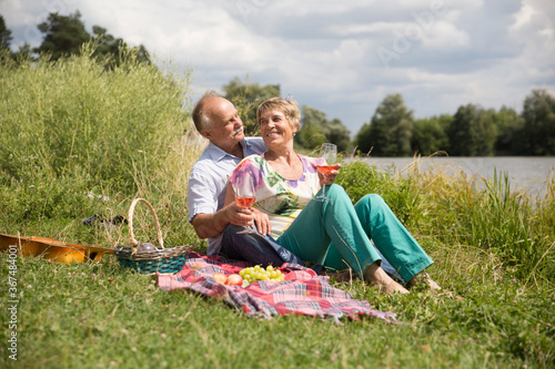  lovely senior couple  having a picnic, drink rose wine on the shore of the lake on a summer day. Staycations, hyper-local travel,  family outing, getaway.  © ulza