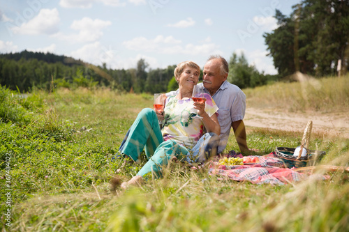  lovely senior couple having a picnic, drink rose wine on the shore of the lake on a summer day. Staycations, hyper-local travel, family outing, getaway. 