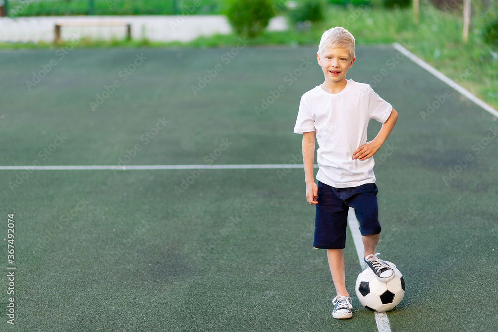 a blond boy in a sports uniform stands on a football field with a soccer ball, sports section. Training of children, children's leisure