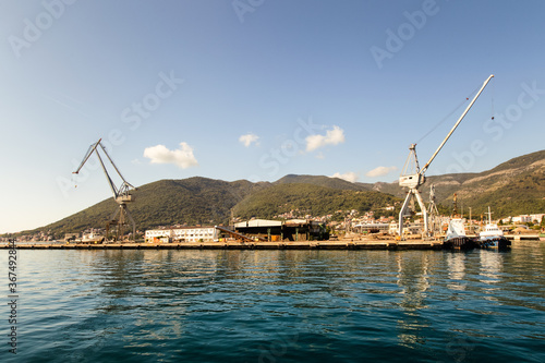 Cranes at cargo terminal in Kotor bay. Montenegro docking station with cranes and tug boats. Mountains, town, blue sky as background.