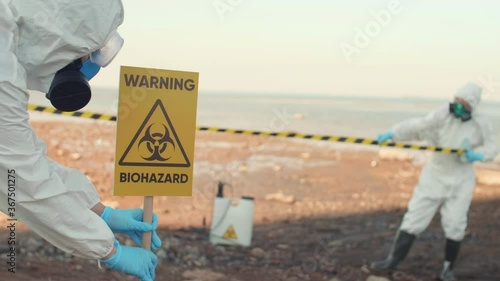 Panning of two scientists wearing chemical suits cordoning off territory where environmental collapse has happened. Scientist installing warning sign and rolling out warning tape photo