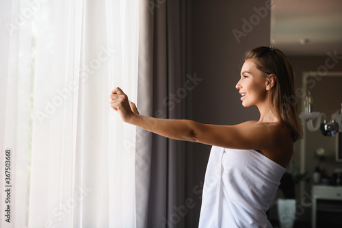 Beautiful woman wearing white towel after shower, open window cover curtains to see morning sun light, enjoy moment at luxury hotel.