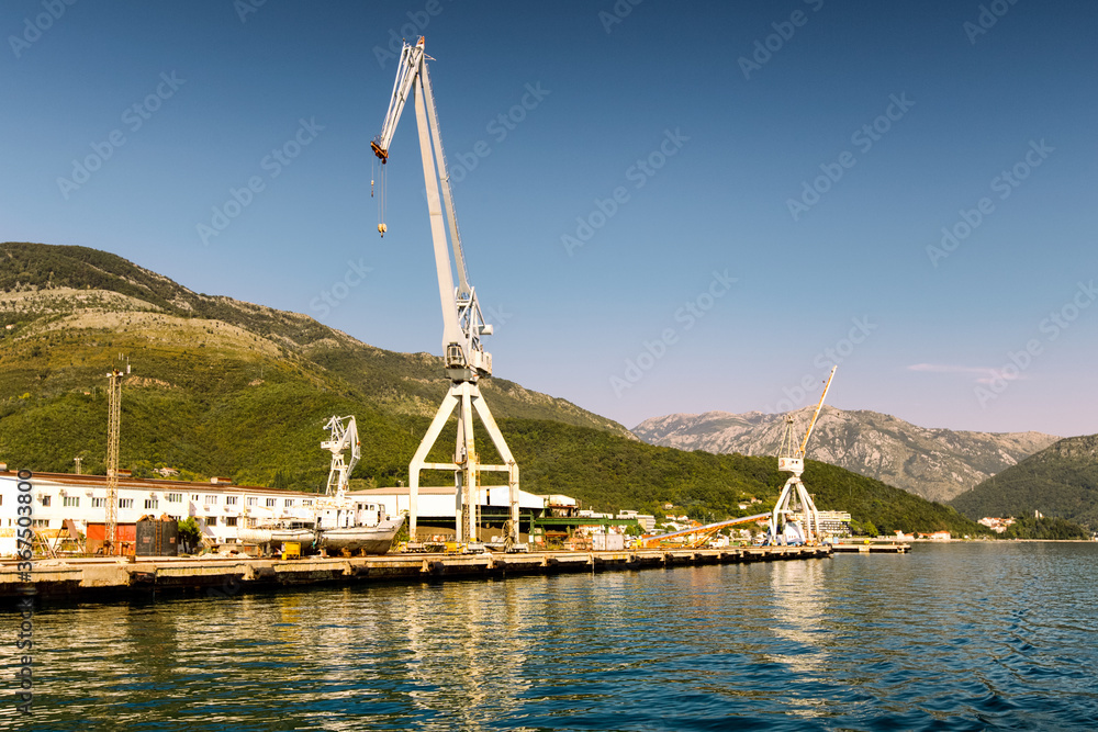 Cranes at cargo terminal in Kotor bay. Montenegro sea dock with cranes and boats. Mountains, town, blue sky as background.