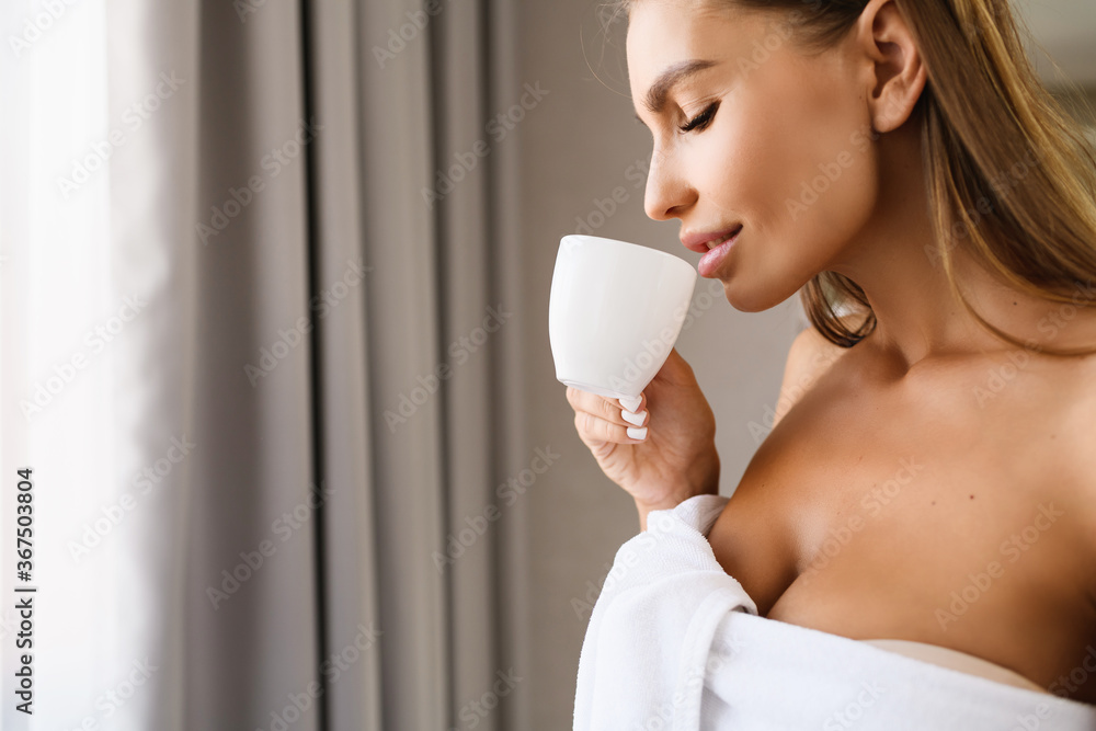 Beautiful young woman with soft skin near window with cup of tea, coffee after shower, wearing white towel, with gray curtains on background. Face of sensual blonde female at happy morning. Beautiful young woman with soft skin near window with cup of tea, coffee after shower, wearing white towel, with gray curtains on background. Face of sensual blonde female at happy morning.