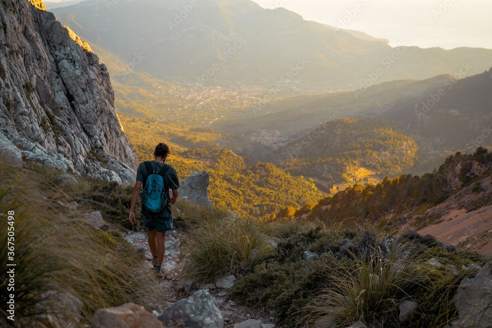 Young spanish man walking through a hiking path in Serra de Tramuntana at golden hour with the village of Soller at the backgorund tinted by golden light