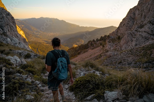 Young spanish man walking through a hiking path in Serra de Tramuntana at golden hour with the village of Soller at the backgorund tinted by golden light