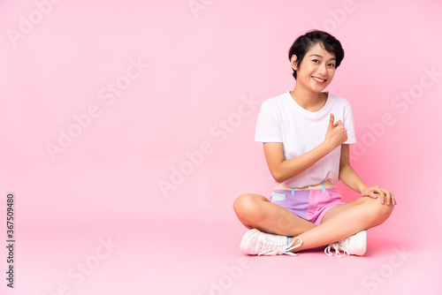 Young Vietnamese woman with short hair sitting on the floor over isolated pink background giving a thumbs up gesture