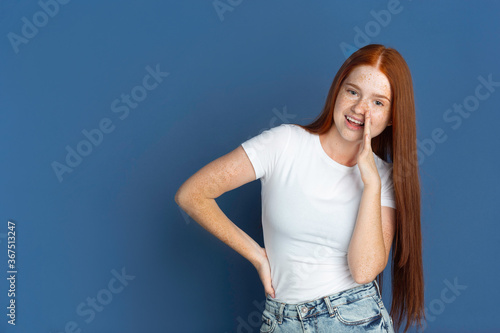 Whispering a secret. Caucasian young girl's portrait on blue background. Beautiful female redhair model with cute freckles. Concept of human emotions, facial expression, sales, ad, youth culture.