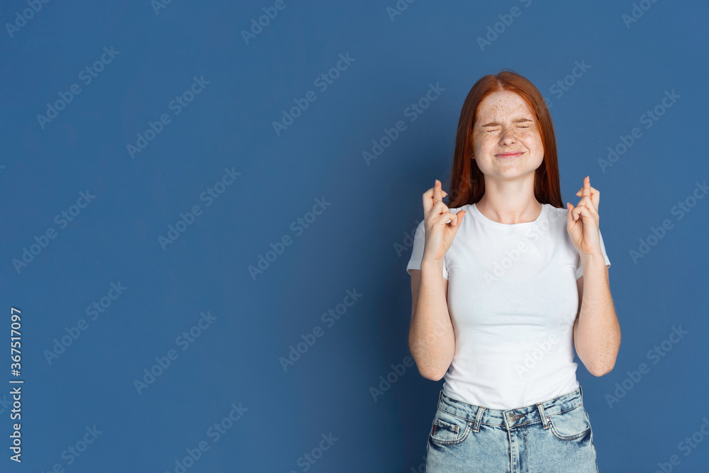 Fingers crossed, hopeful. Caucasian young girl's portrait on blue background. Beautiful female redhair model with cute freckles. Concept of human emotions, facial expression, sales, ad, youth culture. Fingers crossed, hopeful. Caucasian young girl's portrait on blue background. Beautiful female redhair model with cute freckles. Concept of human emotions, facial expression, sales, ad, youth culture.
