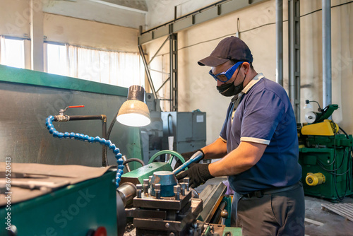 A working turner in an antiviral mask works on a lathe during quarantine. Prevention of the spread of covid 19 within the plant. Worker safety.