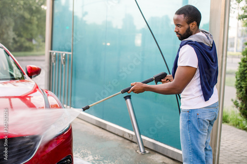 Side view of African handsome young man in casual wear, cleaning his luxury red automobile with high pressure water jet at self car wash service outdoors in summer morning
