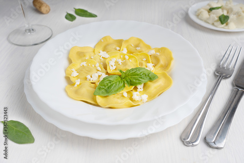 Plate full of italian ravioli pasta filled with cheese decorated by ricotta and basil leaves surrounded by knife and fork, glass of white wine and spinach on white wooden background ready for eating