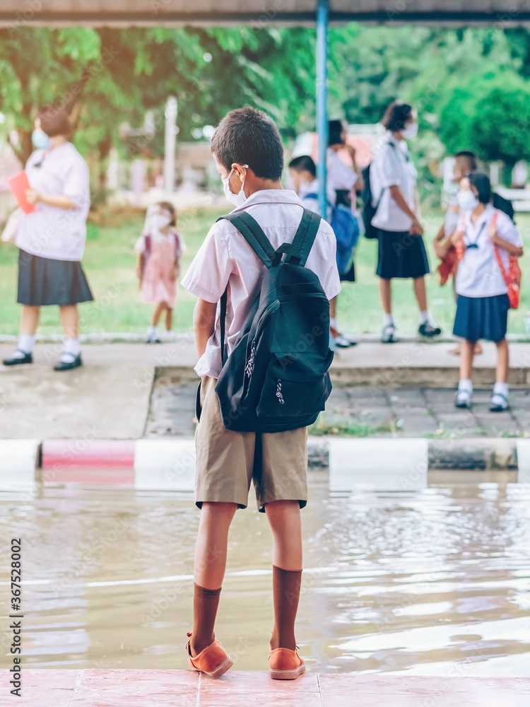 Male elementary school student wear face mask to prevent the Coronavirus(Covid-19) wait for her parents to pick her up to return home after school and the rain just stop in front of the school gate