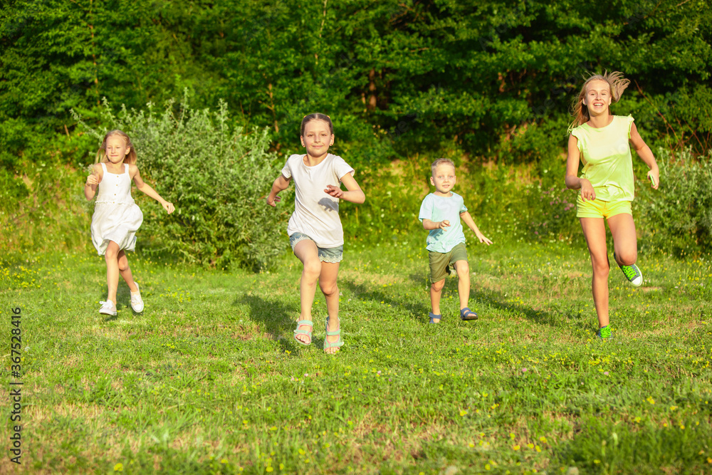 Kids, children running on meadow in summer's sunlight. Look happy, cheerful with sincere bright emotions. Cute caucasian boys and girls. Concept of childhood, happiness, movement, family and summer.