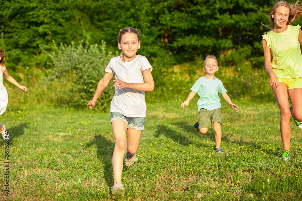 Obraz premium Kids, children running on meadow in summer's sunlight. Look happy, cheerful with sincere bright emotions. Cute caucasian boys and girls. Concept of childhood, happiness, movement, family and summer.